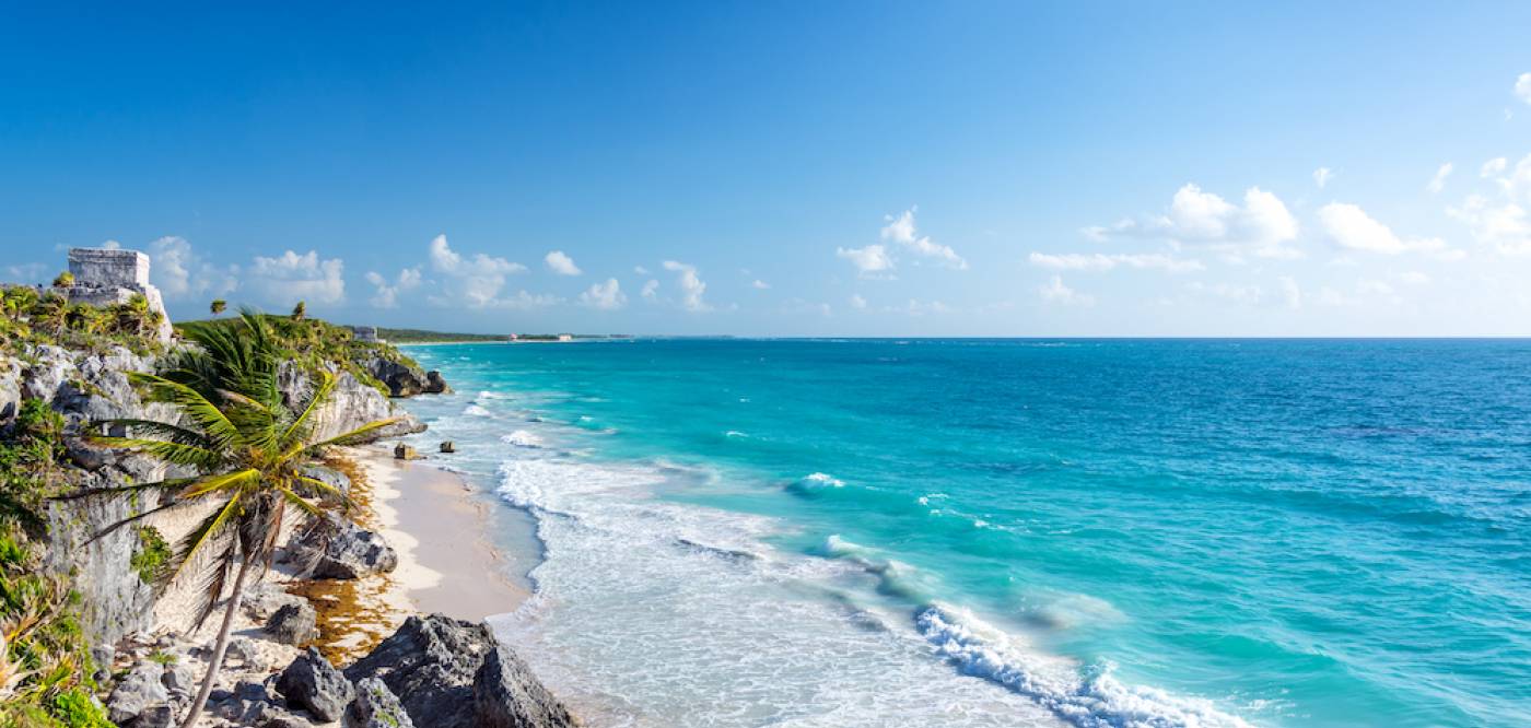 The Tulum ruins sit in the distance along the beach, with waves crashing along the white sandy beaches.